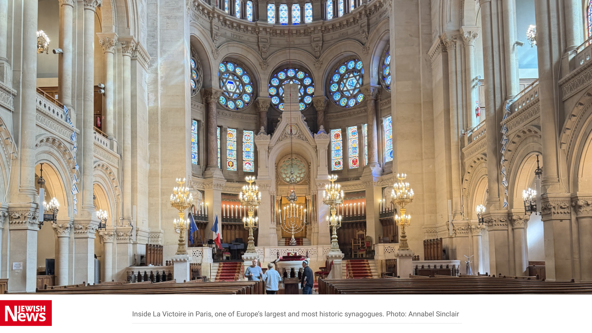 Interior of La Victoire synagogue in Paris with stained-glass windows, chandeliers, columns, and a central menorah.