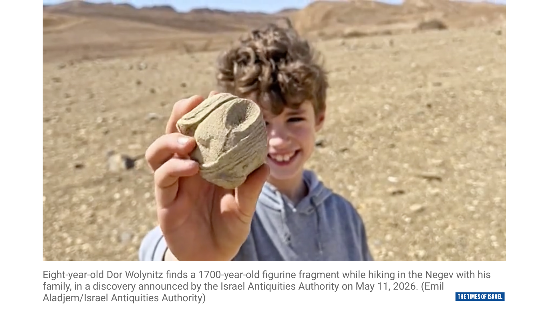 A smiling boy holds a small ancient statuette fragment toward the camera in a desert landscape.