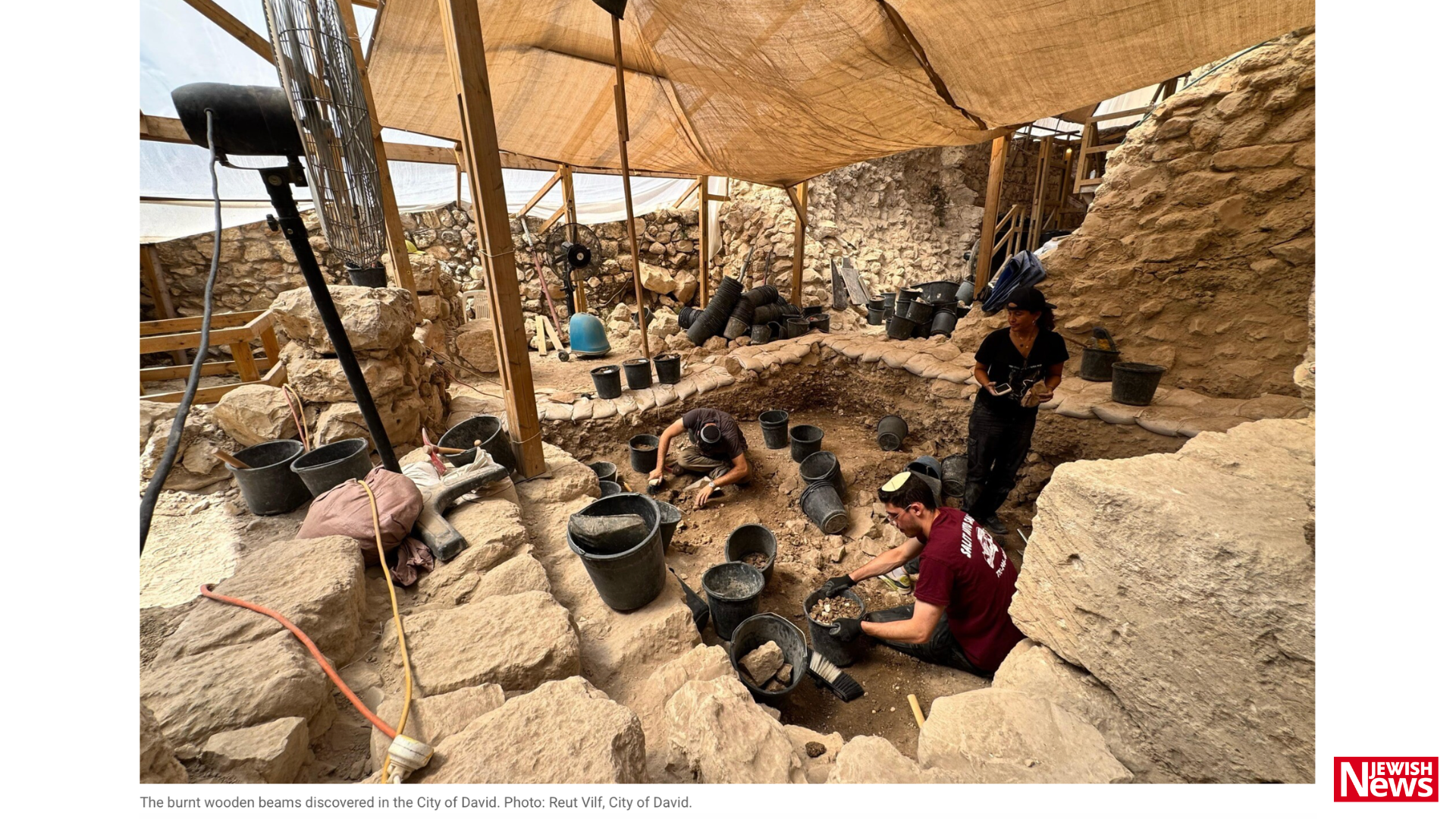 Three people work in a stone-lined archaeological excavation beneath tan shade canopies, surrounded by black buckets and tools.